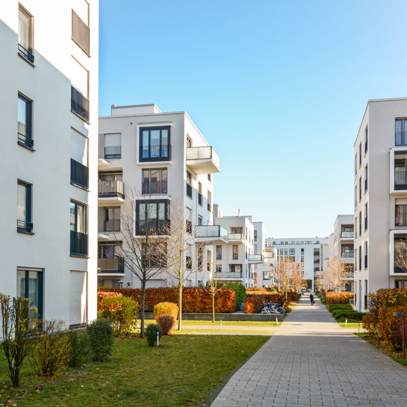 A modern residential complex with white multi-story buildings on either side of a paved walkway. There are neatly maintained green lawns, shrubs, and young trees along the pathway. The sky is clear and blue.