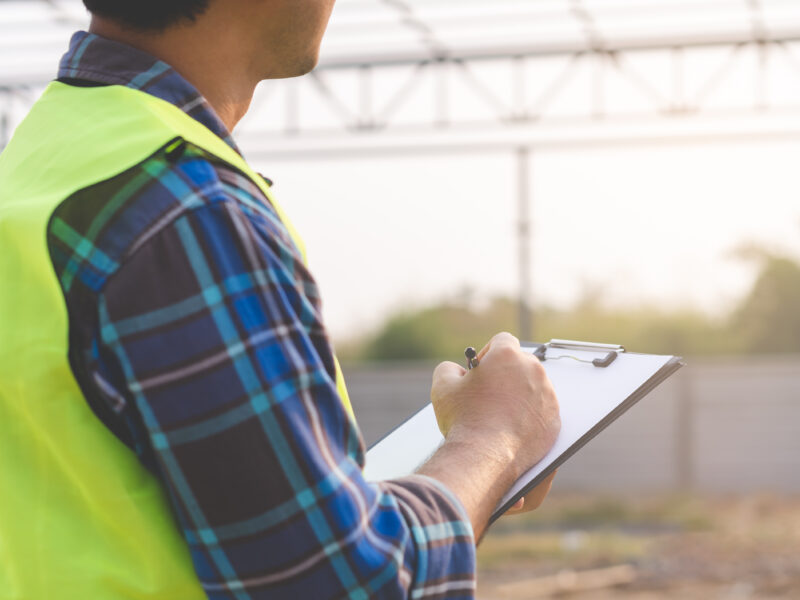 A person in a safety vest holds a clipboard and appears to be writing. They are standing at a construction site with a steel framework structure in the background, and the scene is lit by natural daylight.