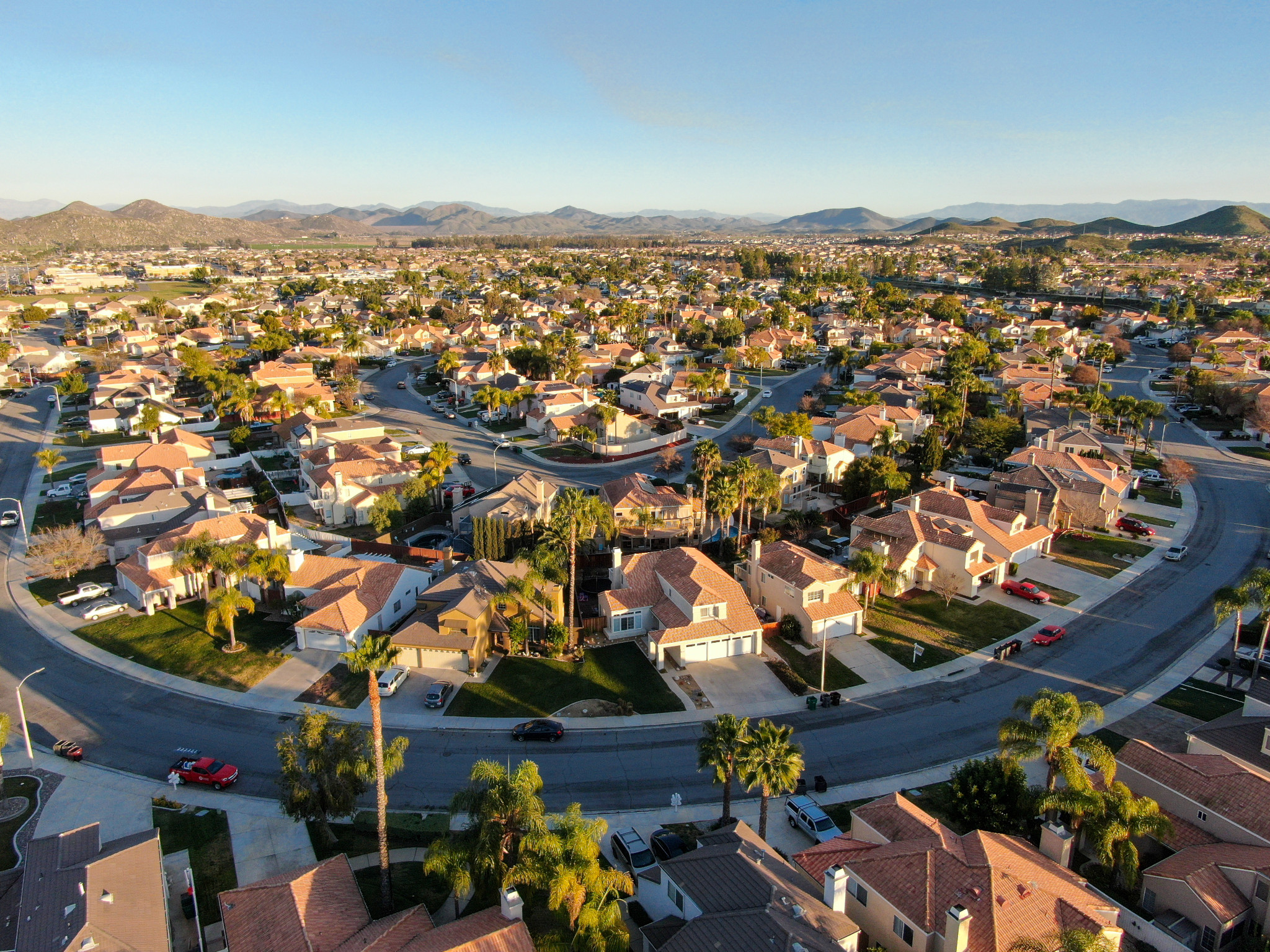 Aerial view of a suburban neighborhood with rows of houses featuring red-tiled roofs, surrounded by curved streets. The landscape is dotted with trees, and mountains are visible in the background under a clear blue sky.