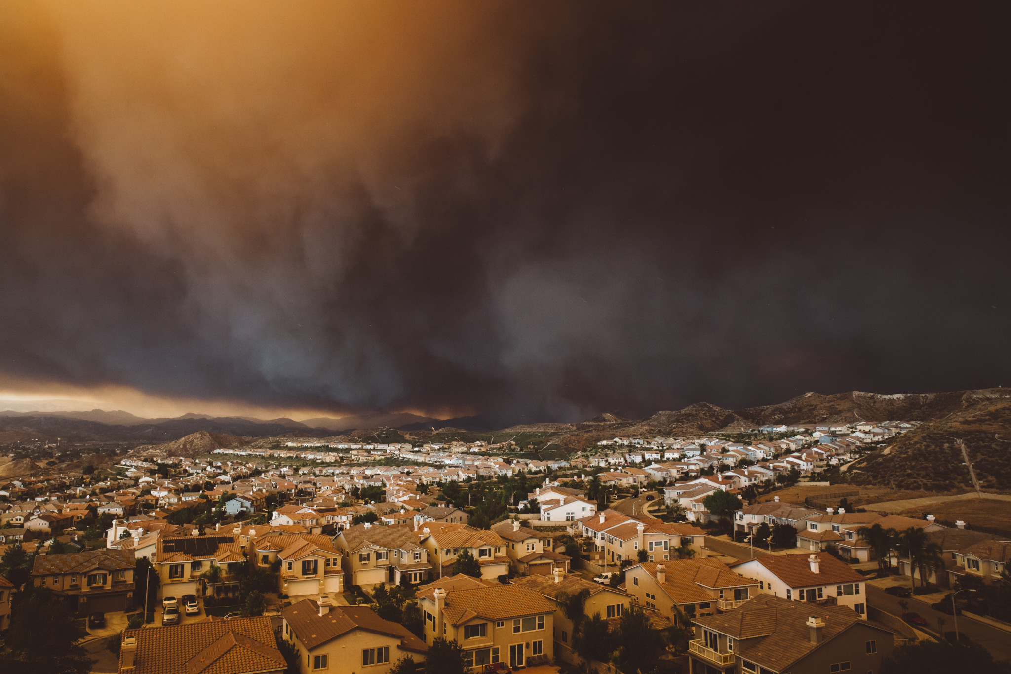 A suburban neighborhood under a dark, smoky sky from a nearby wildfire. The orange glow from the fire contrasts with the rooftops of densely packed houses and the surrounding hills.