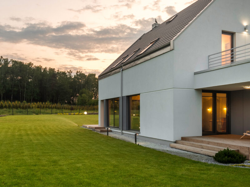 Modern house with sloped roof and large windows at dusk, surrounded by a spacious, well-maintained lawn. A forested area is visible in the background under a partly cloudy sky. Exterior lights illuminate the home's entrance.