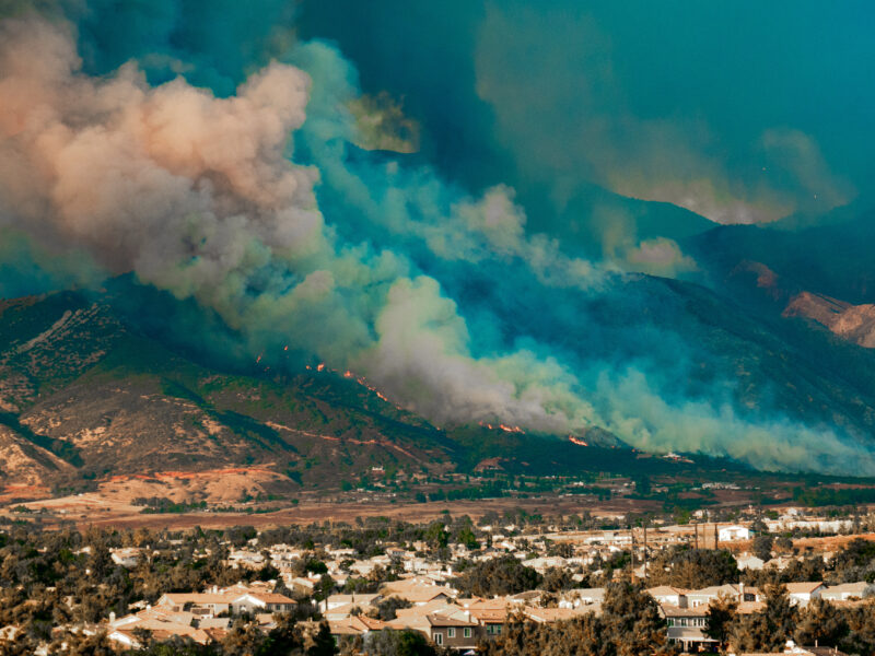 A wildfire spreads across a mountain range near a residential area. Thick plumes of smoke, colored in shades of blue and orange, rise into the sky. The foreground shows houses and trees under a clear blue-green sky.