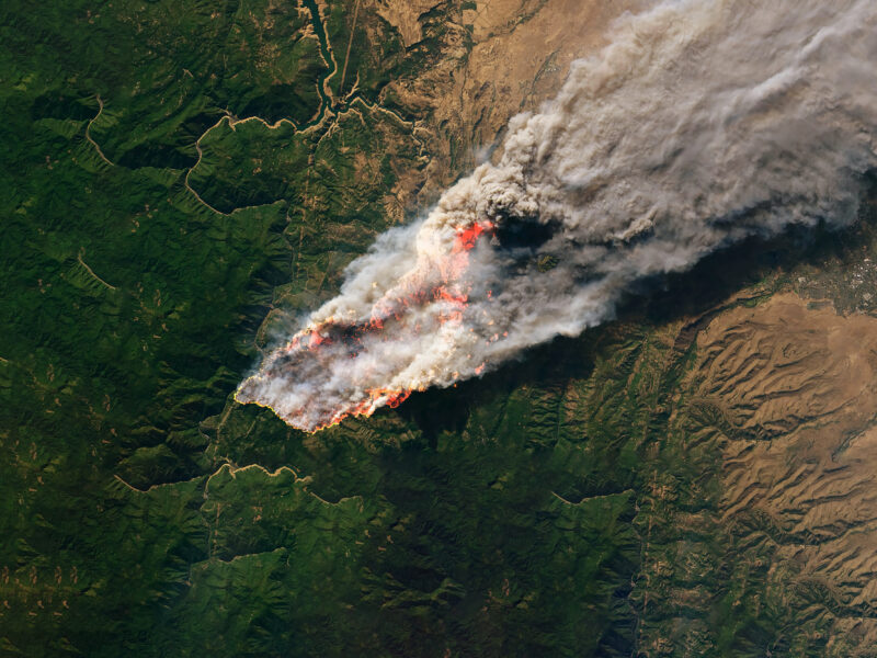 Aerial view of a large wildfire engulfing a forested region, with thick smoke billowing into the sky. The flames are spreading across green terrain, leaving scorched earth in their path.